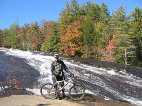 A cyclist stands beside a mountain bike on a rocky surface near a waterfall, surrounded by vibrant fall foliage in trees. The clear blue sky adds to the scenic outdoor setting. DuPont State Recreational Forest mountain bike trail.