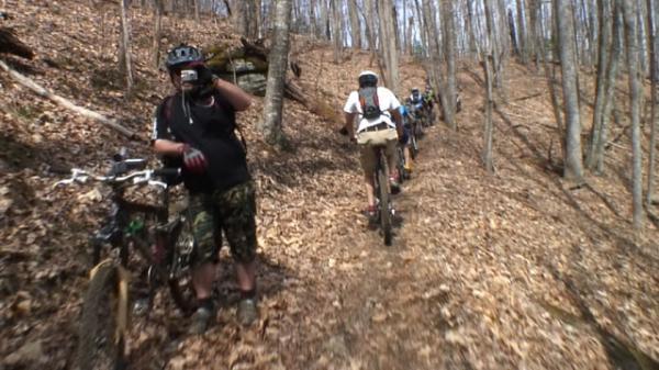 A group of mountain bikers riding along a narrow trail through a wooded area. One cyclist is stopped, taking a picture with a camera, while others continue to ride. The ground is covered with fallen leaves, and trees are bare, indicating early spring. DuPont State Forest mountain bike trail.