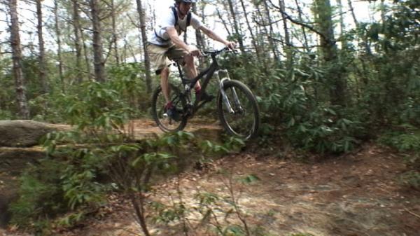 A mountain biker jumping off a natural ramp on a wooded trail, surrounded by trees and underbrush. DuPont State Recreational Forest mountain bike trail.
