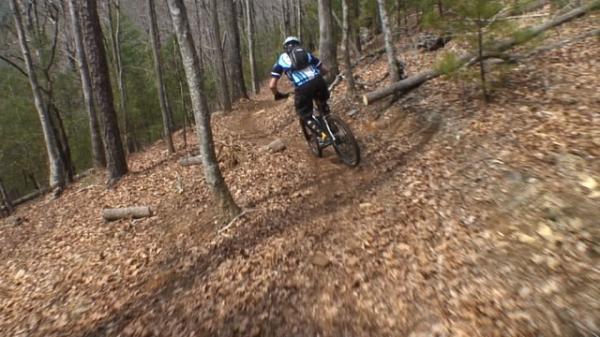A mountain biker navigating a winding trail through a forest, with fallen leaves covering the ground and trees surrounding the path. DuPont State Recreational Forest mountain bike trail.
