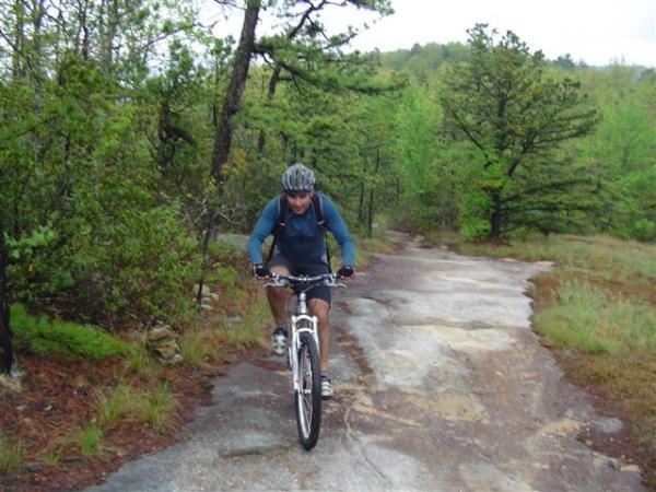 A mountain biker navigating a rocky trail surrounded by lush green trees and vegetation on a cloudy day. The cyclist is wearing a helmet and backpack, focused on riding through the natural terrain. DuPont State Forest mountain bike trail.