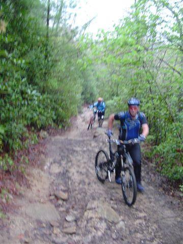 A cyclist navigating a rocky, narrow trail through a dense forest, with another cyclist visible in the background. Both riders are wearing helmets and cycling gear. The scene features greenery and a rugged path typical of mountain biking trails. DuPont State Forest mountain bike trail.