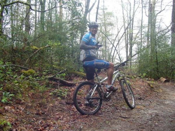 A mountain biker in a blue and gray cycling jersey and helmet, giving a thumbs-up while sitting on a bicycle on a dirt trail surrounded by trees and greenery. DuPont State Recreational Forest mountain bike trail.