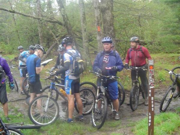 A group of mountain bikers gathered on a muddy trail, consulting a map and discussing their route. They are wearing helmets and cycling gear, with various types of mountain bikes parked nearby. The scene features a forested background with trees and a trail marker labeled "Beau's Loop." DuPont State Recreational Forest mountain bike trail.