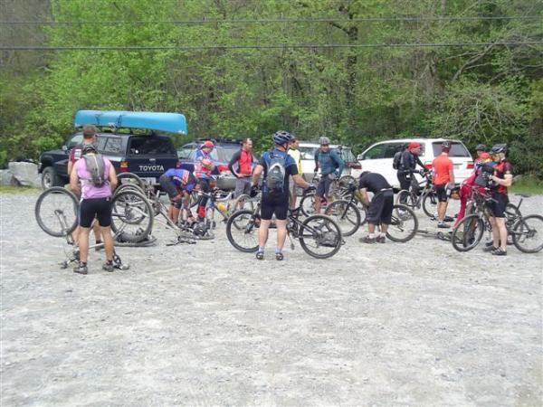 A group of mountain bikers gathered in a gravel parking area, working on their bicycles. Some riders are adjusting their bikes, while others are socializing or preparing for a ride. In the background, a couple of vehicles are parked, including one with a canoe on top. Lush greenery surrounds the scene, indicating a natural outdoor setting. DuPont State Recreational Forest mountain bike trail.