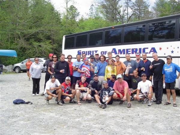 A diverse group of people gathered outdoors, standing together in front of a bus labeled "Spirit of America." The setting appears to be a recreational area, possibly a campsite. Participants are wearing casual clothing, some in sports attire, and are posing with smiles. The group includes both men and women, with various ages represented. In the background, trees and parked vehicles can be seen, suggesting a nature-related activity or event. DuPont State Recreational Forest mountain bike trail.
