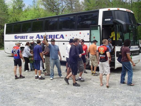 A group of people gathers around a white charter bus labeled "Spirit of America." The scene takes place outdoors in a forested area, with trees in the background. Individuals are interacting as some prepare to board the bus, while others appear to be socializing. The weather looks clear and bright, suggesting a pleasant day. DuPont State Forest mountain bike trail.