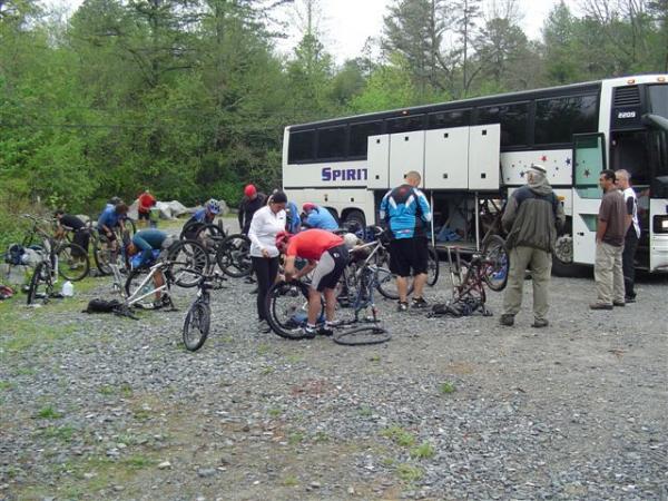 A group of cyclists is gathered near a large bus in a gravel area surrounded by trees. Some individuals are busy repairing their bicycles, while others are conversing. The scene is set outdoors, reflecting a casual, collaborative atmosphere as the cyclists prepare for their ride. DuPont State Forest mountain bike trail.