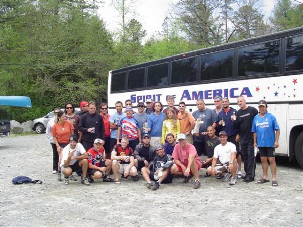 A group of approximately 25 people posing for a photo in front of a large bus labeled "Spirit of America." They are standing on gravel, surrounded by trees and greenery, wearing casual clothing suitable for outdoor activities. Everyone appears to be in good spirits, some holding drinks, and the setting looks like a recreational area. DuPont State Forest mountain bike trail.