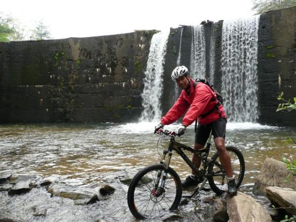 A person in a red jacket and helmet is riding a mountain bike near a waterfall, surrounded by rocks and water. The scene captures the natural beauty of the area, with the cascading water in the background and greenery visible in the surroundings. DuPont State Recreational Forest mountain bike trail.