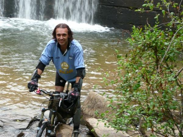 A man in a light blue cycling jersey and black shorts stands beside his mountain bike in a creek, with a waterfall in the background. He is wearing protective gloves and smiling at the camera, surrounded by greenery. DuPont State Recreational Forest mountain bike trail.