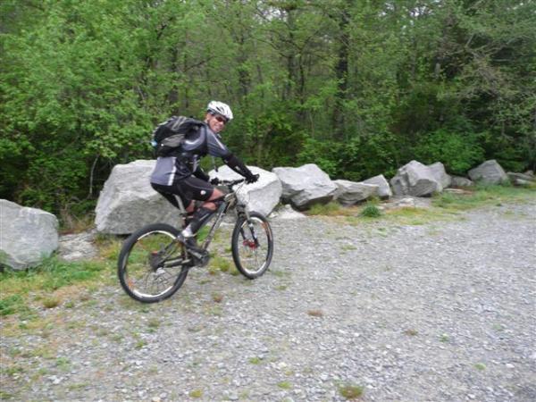 A person in sportswear riding a mountain bike along a gravel path surrounded by greenery and large rocks. The individual is wearing a helmet and carrying a backpack, with trees and foliage in the background. DuPont State Forest mountain bike trail.
