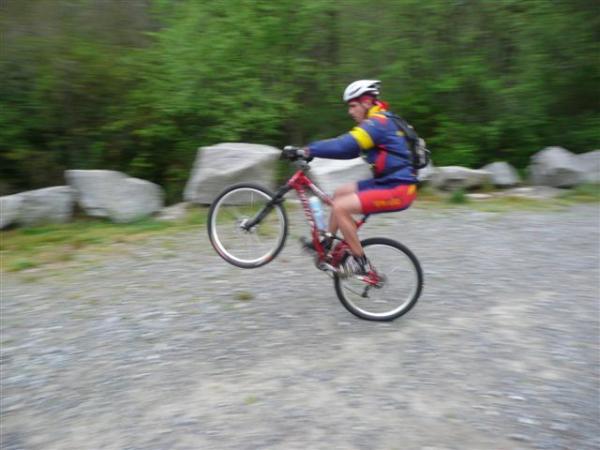 A mountain biker performing a wheelie on a gravel path, surrounded by large rocks and greenery. The cyclist is wearing a helmet and colorful riding gear, showcasing action and skill. DuPont State Forest mountain bike trail.