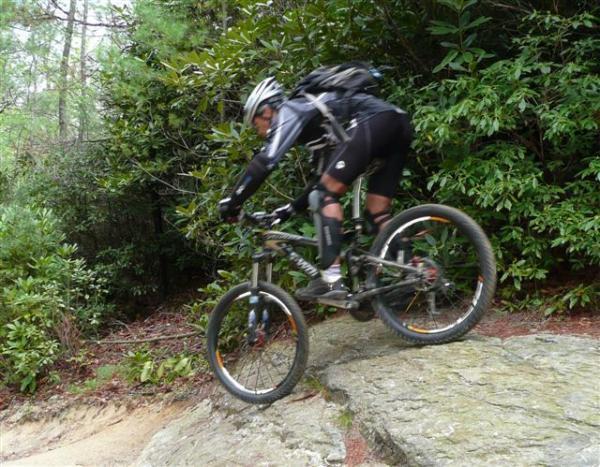 A mountain biker navigating rocky terrain, captured in mid-air while descending from a large rock. The cyclist is wearing a helmet and protective gear, surrounded by lush greenery in a natural outdoor setting. DuPont State Forest mountain bike trail.
