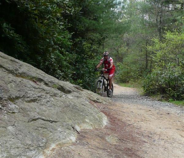 A cyclist riding a mountain bike on a dirt trail surrounded by trees and rocks. The path winds through a wooded area, with lush greenery on either side and a prominent rock formation nearby. DuPont State Recreational Forest mountain bike trail.