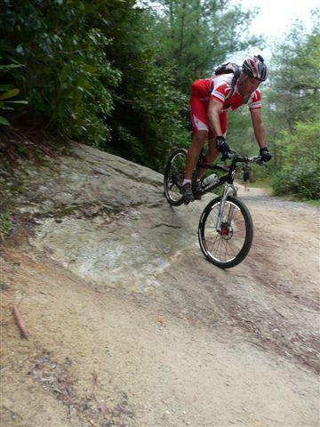 A cyclist in a red and white outfit skillfully navigates down a rocky, sloped trail amidst lush greenery, showcasing an adventurous mountain biking scene. DuPont State Forest mountain bike trail.