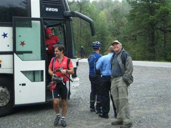 A group of four men near a parked tour bus in a wooded area. One man in cycling attire gets off the bus, while another in casual clothes stands outside smiling. Two other men are conversing by the bus. The setting appears to be a recreational stop with greenery in the background. DuPont State Recreational Forest mountain bike trail.