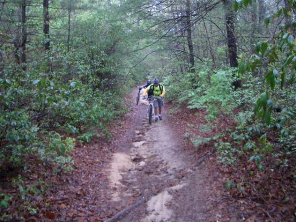 A narrow dirt trail winding through a lush, green forest with trees and bushes on either side. Two individuals are seen walking along the path, one carrying a bicycle. The ground appears slightly muddy, suggesting recent rainfall. DuPont State Recreational Forest mountain bike trail.