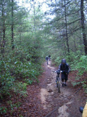 Two mountain bikers navigate a narrow, muddy trail surrounded by dense trees and greenery. The scene captures a lush, natural environment with damp soil and foliage, suggesting a rainy setting. DuPont State Recreational Forest mountain bike trail.