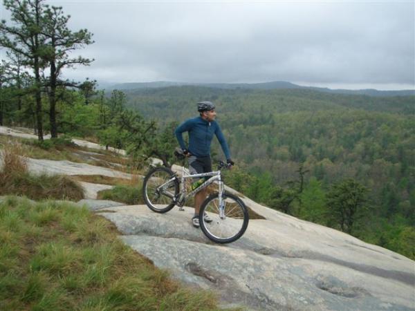 A mountain biker standing on rocky terrain, wearing a blue long-sleeve shirt and helmet, overlooking a green valley and distant hills under a cloudy sky. DuPont State Recreational Forest mountain bike trail.