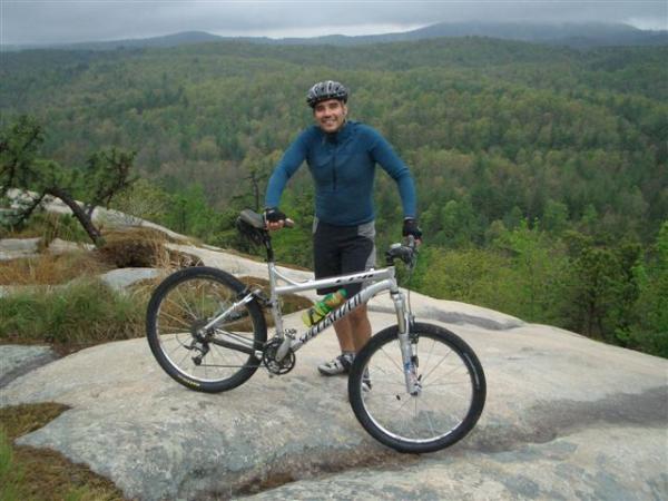 A person smiling while standing next to a mountain bike on a rocky outcrop, with a lush green forest and mountains in the background under overcast skies. DuPont State Forest mountain bike trail.