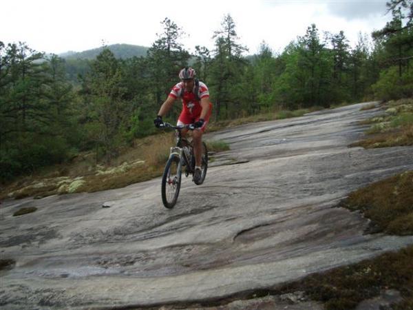 A mountain biker in a red and white jersey and helmet jumps over a rocky section of trail, surrounded by trees and a hilly landscape. DuPont State Forest mountain bike trail.