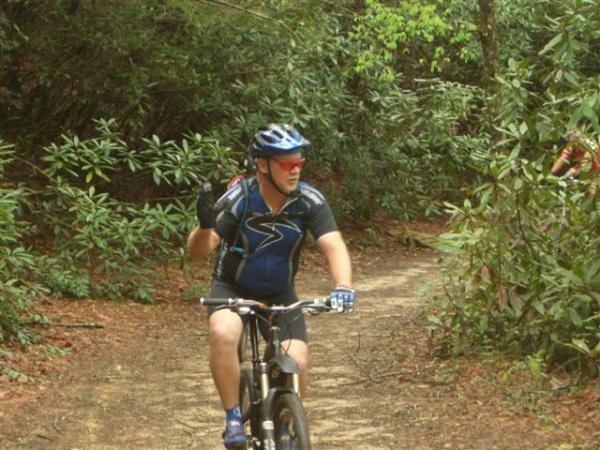 A person riding a mountain bike on a dirt trail surrounded by greenery, wearing a blue helmet and a short-sleeved cycling jersey. They are gesturing with one hand while pedaling. DuPont State Recreational Forest mountain bike trail.