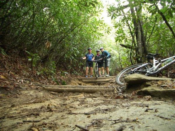 Two mountain bikers are standing on a rough, muddy trail surrounded by dense greenery in a forested area. A bicycle lies on its side nearby, and the path is uneven with exposed roots and rocks. The scene captures the adventurous spirit of mountain biking in a natural setting. DuPont State Recreational Forest mountain bike trail.