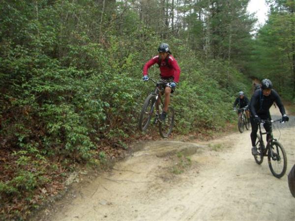 A group of mountain bikers navigating a dirt trail in a wooded area. One cyclist, wearing a red jacket and helmet, is performing a jump over a small mound, while two other riders follow closely behind on the path. Lush greenery surrounds the trail, creating a natural setting for outdoor cycling. DuPont State Forest mountain bike trail.