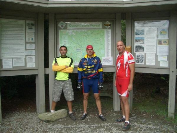 Three cyclists stand in front of a map and information display for Dupont State Forest. The first cyclist, on the left, is wearing a bright yellow shirt and gray shorts. The second, in the center, is dressed in a blue and yellow cycling outfit with a red bandana. The third cyclist, on the right, is in a red and white cycling jersey. Surrounding them are trees and informational panels about the forest. DuPont State Recreational Forest mountain bike trail.