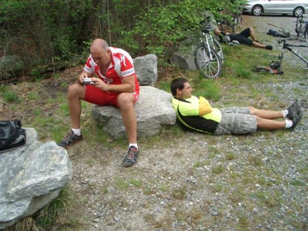 Two men resting on rocks in a natural setting after cycling. One man, dressed in a red and white cycling outfit, sits on a rock, using a device, while the other man, wearing a yellow and black shirt, reclines on the ground nearby. Bicycles are visible in the background, along with trees and a gravel path. DuPont State Recreational Forest mountain bike trail.