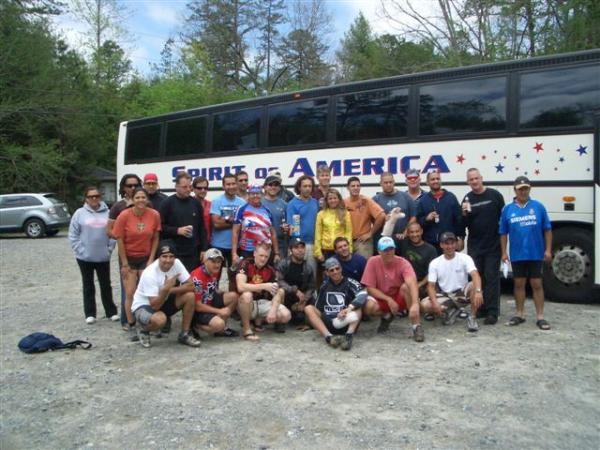 A group of diverse individuals standing together in front of a bus labeled "Spirit of America." The scene takes place outdoors, amidst trees and gravel, on a sunny day. The group is smiling and wearing casual clothing, with some individuals seated on the ground and others standing. DuPont State Forest mountain bike trail.