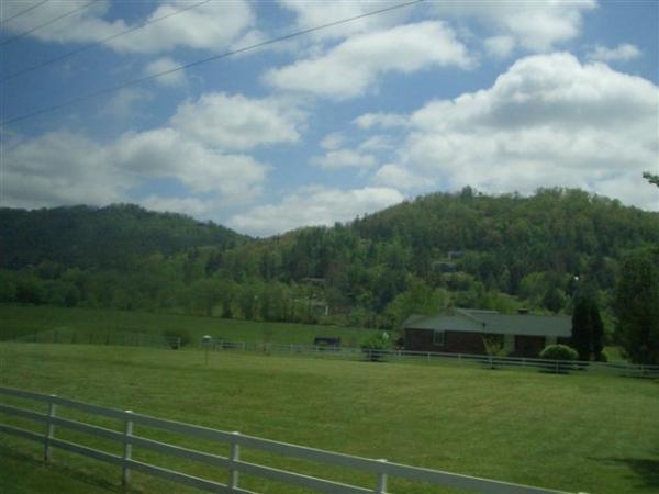 A scenic landscape featuring rolling green hills under a partly cloudy sky. In the foreground, a lush green field bordered by a white fence leads to a rural building, surrounded by trees. The hills in the background are densely wooded, showcasing various shades of green. DuPont State Forest mountain bike trail.