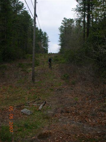 A cyclist riding on a narrow, overgrown trail surrounded by trees. Power lines are visible in the background, and the scene is set in a natural, wooded area during cloudy weather. The ground is covered with leaves and small debris. DuPont State Forest mountain bike trail.