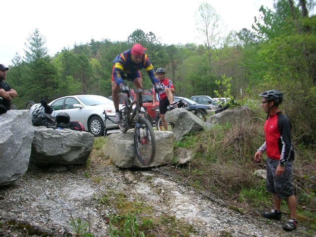 A mountain biker in a colorful jersey leaps over a large rock while other cyclists observe nearby. The scene is set in a wooded area with parked cars in the background, surrounded by greenery. DuPont State Recreational Forest mountain bike trail.
