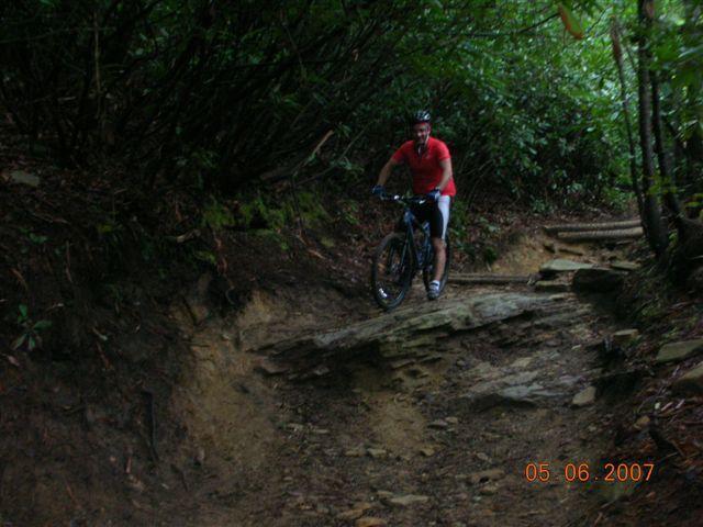 A mountain biker in a red shirt navigates a rocky trail surrounded by dense greenery. The rider is on a black bike, positioned on a section of uneven terrain, demonstrating skill and balance amidst the natural landscape. DuPont State Forest mountain bike trail.