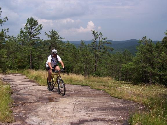 A mountain biker rides on a rocky trail surrounded by green trees and hills under a cloudy sky. DuPont State Recreational Forest mountain bike trail.