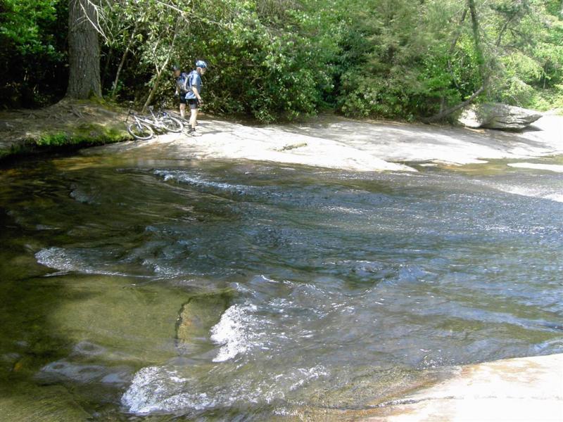 A person standing beside a flowing stream in a wooded area, with a mountain bike resting on the ground nearby. The scene features smooth rock surfaces and lush greenery in the background, reflecting a serene outdoor setting. DuPont State Forest mountain bike trail.