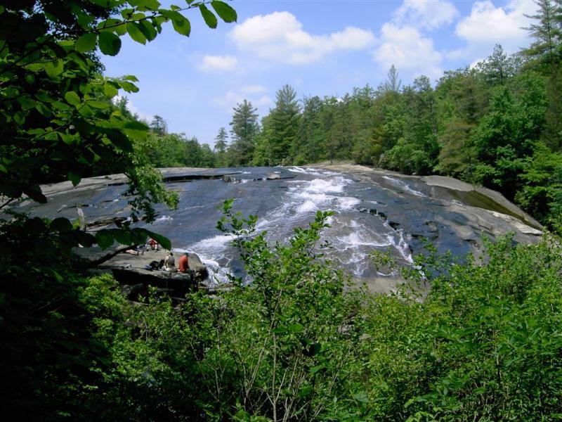 A scenic view of a rocky hillside with smooth, dark stones partially covered by flowing water, surrounded by lush green foliage and trees under a blue sky with fluffy clouds. DuPont State Forest mountain bike trail.