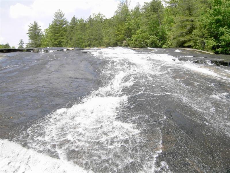 A smooth, rocky surface with water flowing over it, surrounded by lush green trees under a partly cloudy sky. DuPont State Recreational Forest mountain bike trail.