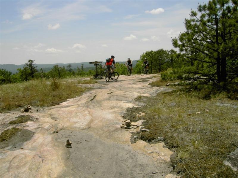 Three mountain bikers navigate a rocky trail atop a hillside surrounded by trees and distant mountains under a partly cloudy sky. Small stone cairns are visible along the path, indicating a hiking route. DuPont State Forest mountain bike trail.
