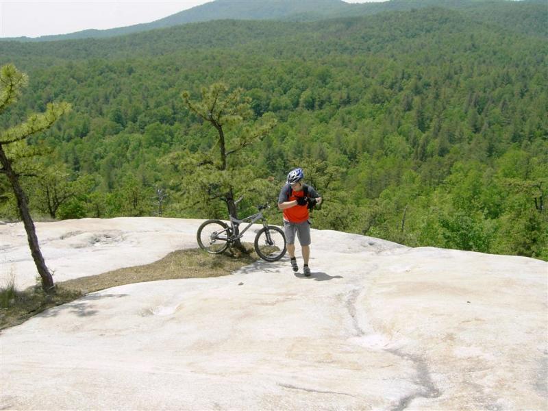 A mountain biker in a gray helmet and orange shirt walks on a rocky outcrop, carrying a camera. A mountain bike rests nearby, surrounded by lush trees and rolling hills in the background under a clear sky. DuPont State Forest mountain bike trail.
