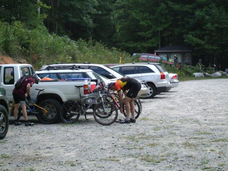 Two cyclists are preparing their mountain bikes in a gravel parking area, surrounded by parked cars and lush greenery. One cyclist, wearing a yellow and red jersey, is adjusting their bike, while the other, in a darker outfit, is handling theirs. In the background, there are additional vehicles and a small structure, suggesting a recreational area. DuPont State Forest mountain bike trail.