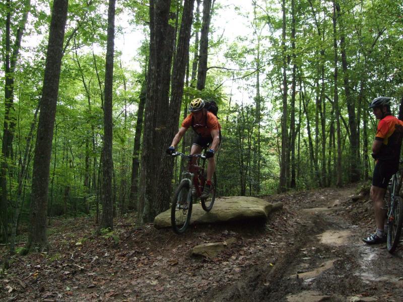 Mountain biker in an orange shirt and helmet skillfully navigating over a rock on a dirt trail, surrounded by lush green trees. Another cyclist in the background observes, wearing a matching shirt and helmet. DuPont State Recreational Forest mountain bike trail.