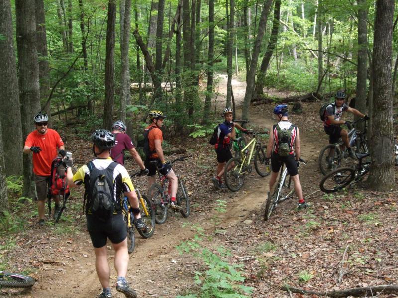 A group of six mountain bikers wearing helmets and athletic gear are gathered on a dirt trail in a lush green forest. Some riders are standing beside their bikes while others are conversing, with trees and foliage surrounding them. The path ahead leads further into the woods. DuPont State Forest mountain bike trail.
