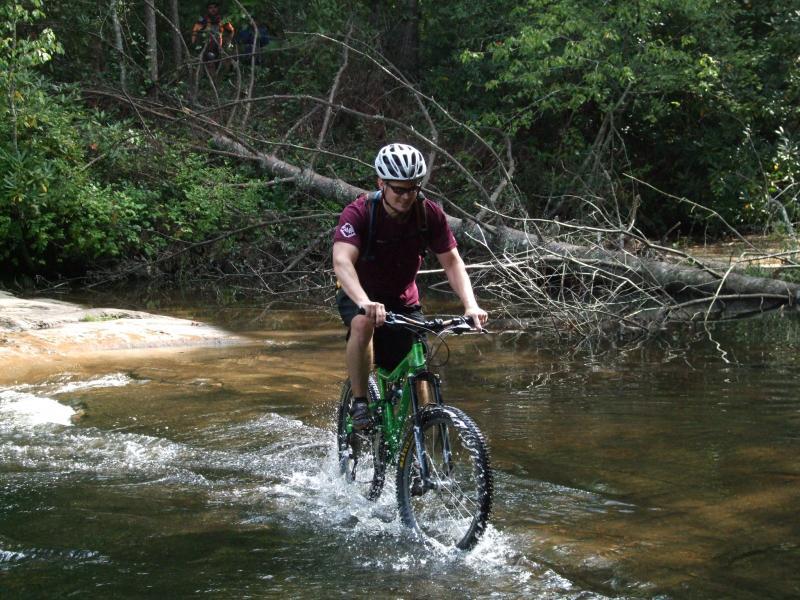 A person riding a green mountain bike through shallow water in a forested area, with a helmet on and a backpack. Trees and a fallen branch can be seen in the background, creating a natural outdoor setting. DuPont State Forest mountain bike trail.