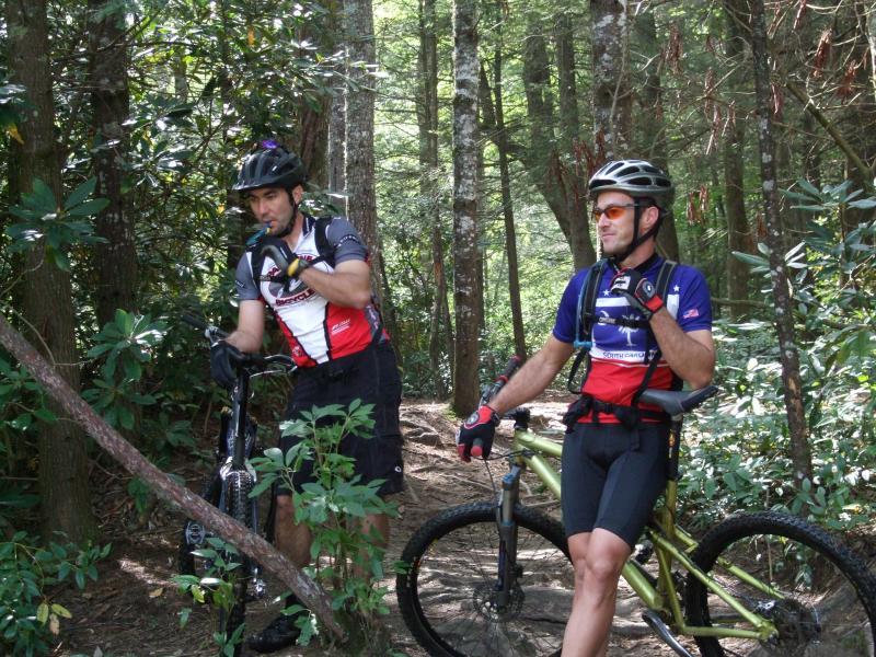 Two mountain bikers pause on a forest trail, one drinking water from a hydration pack and the other standing beside his bike. Both are wearing helmets and cycling attire, surrounded by trees and greenery. DuPont State Recreational Forest mountain bike trail.