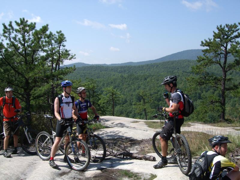 A group of five mountain bikers pause on a rocky overlook, surrounded by lush green trees and rolling hills under a clear blue sky. Two bikers stand with their bikes, engaged in conversation, while the others are positioned nearby, taking in the scenic view. The riders are dressed in cycling gear, showcasing a mix of bright colors and styles. DuPont State Forest mountain bike trail.