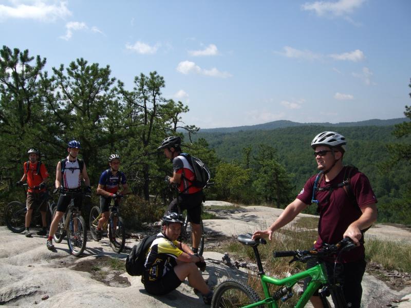 A group of six mountain bikers resting on a rocky outcrop, surrounded by trees and a scenic hilly landscape under a blue sky with fluffy clouds. Some riders are sitting or standing with their bikes, chatting and enjoying the view. DuPont State Forest mountain bike trail.