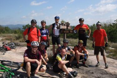 A group of ten mountain bikers poses for a photo in front of a trail sign indicating "Big Rock Trail" and "Rock Trail." The scene is set outdoors on a rocky surface with green trees and hills in the background under a partly cloudy sky. Some bikes are laid on the ground nearby. The group is smiling, dressed in cycling gear and helmets, showcasing their enthusiasm for biking. DuPont State Recreational Forest mountain bike trail.